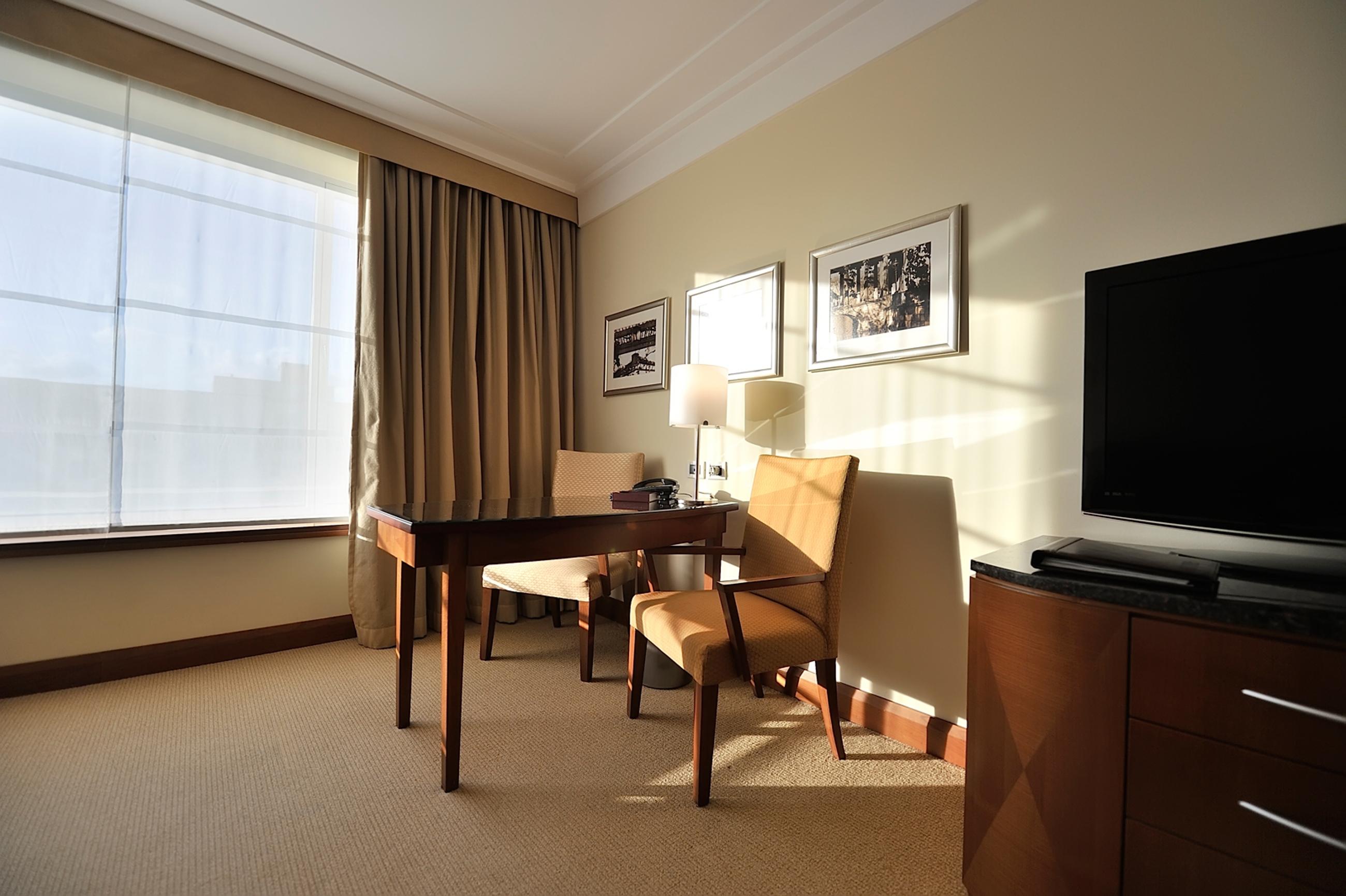 Regent Warsaw Hotel Buitenkant foto The photo shows a well-lit hotel room or office space. On one side, there is a large window with light-colored curtains that allow natural light to enter. Near the window, there are two upholstered chairs, one of which is positioned at a wooden desk.