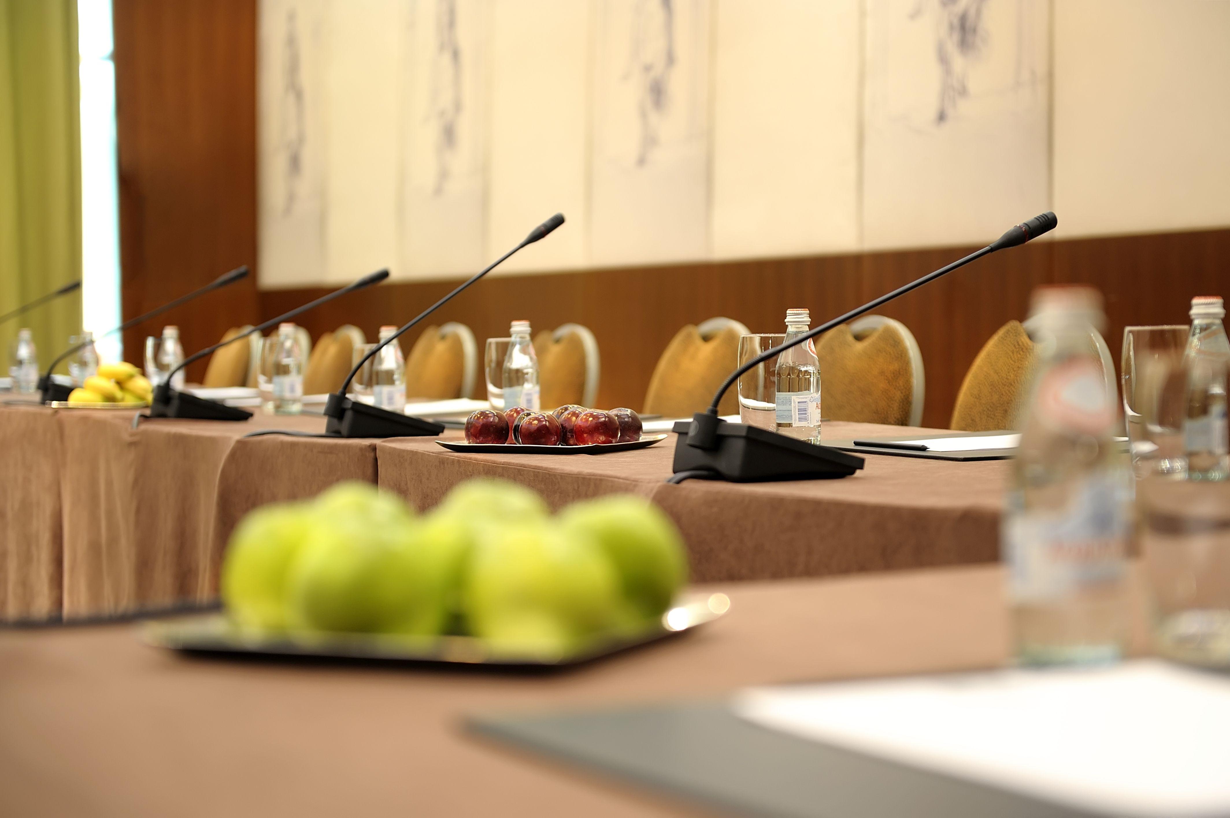Regent Warsaw Hotel Buitenkant foto The photo depicts a conference room setup. There are several chairs arranged around a rectangular table covered with a brown tablecloth. Each place at the table has a microphone and a water bottle in front. On the table, there is a plate of dates and