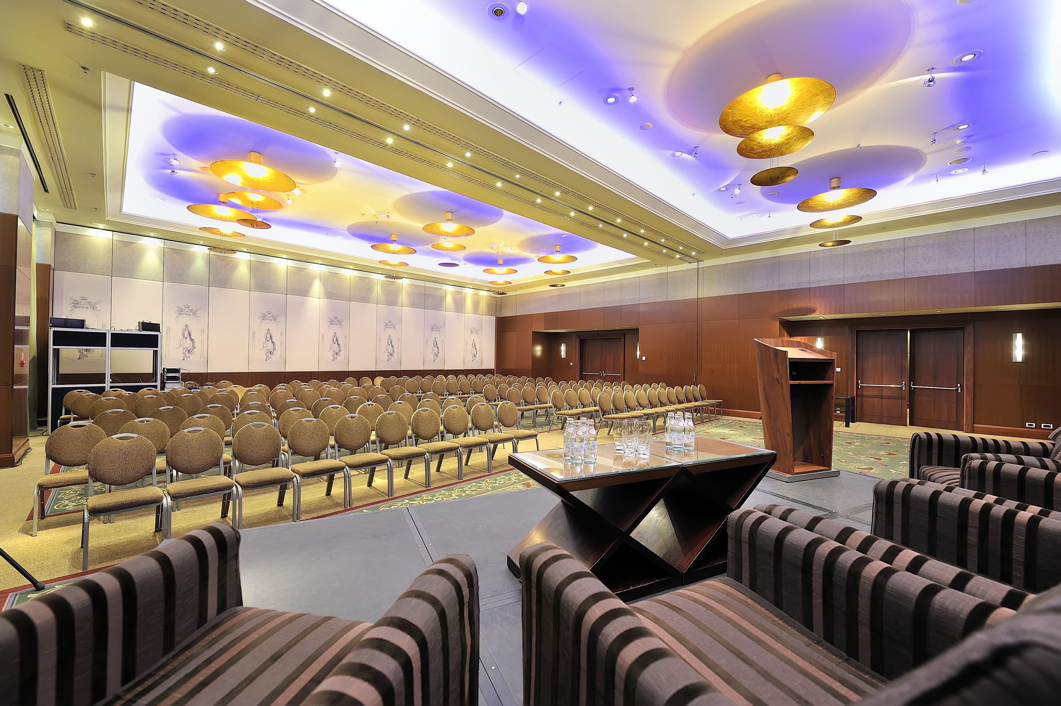 Regent Warsaw Hotel Buitenkant foto The photo shows a conference room or event space set up for a presentation or meeting. There are several rows of beige chairs arranged in front of a podium, which is positioned on a small stage. The room features elegant lighting, with decorative cei