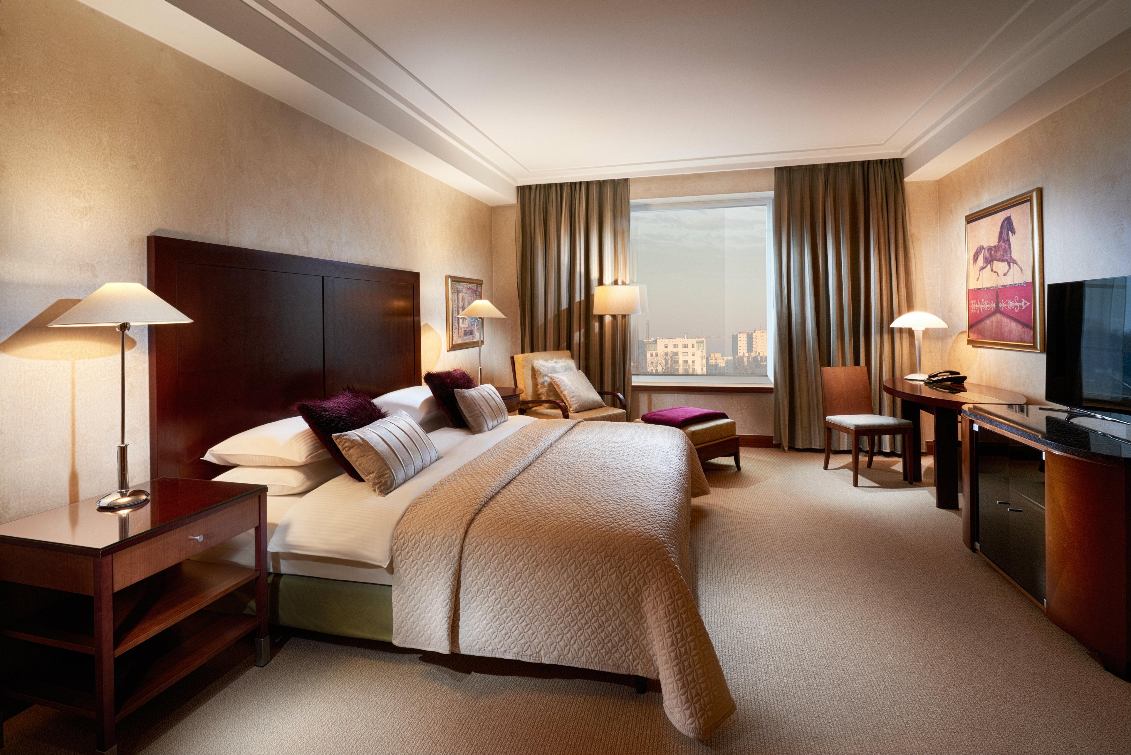 Regent Warsaw Hotel Buitenkant foto The photo shows a well-appointed hotel room featuring a large bed with white and cream bedding. There are bedside lamps on either side of the bed, and a comfortable armchair is positioned nearby. A window with long curtains allows natural light to en