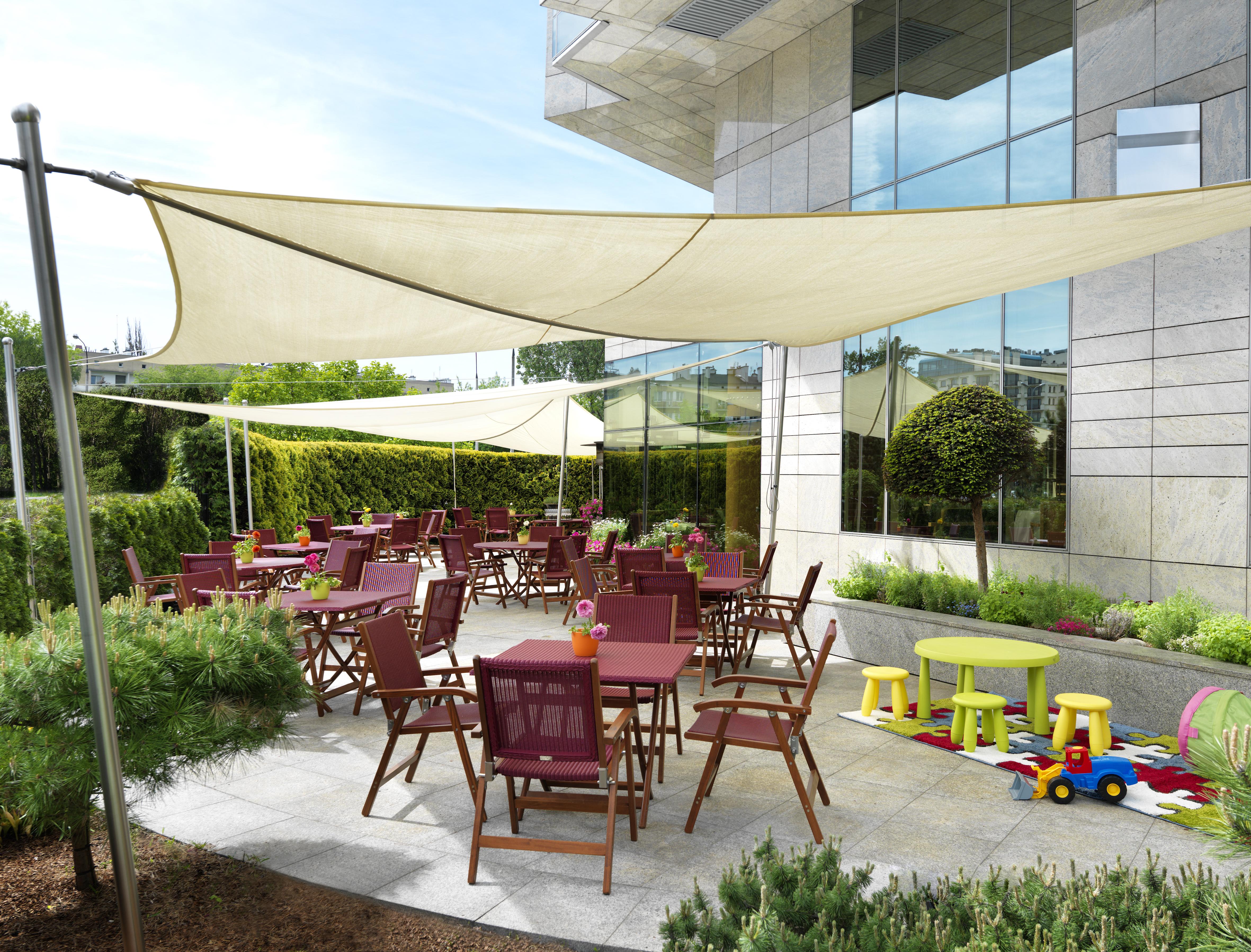Regent Warsaw Hotel Faciliteiten foto The photo shows an outdoor dining area with a modern design. There are several wooden tables and chairs arranged on a paved surface, with some tables set for dining. Above the seating area, there are large fabric canopies providing shade. In addition
