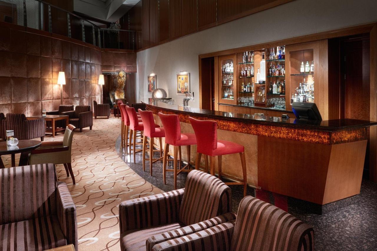 Regent Warsaw Hotel Buitenkant foto The photo shows a stylish and sophisticated bar interior. There are several tall bar stools with red upholstery positioned along a polished wooden bar counter. Behind the bar, shelves display a variety of bottles and glassware, indicating a well-stoc