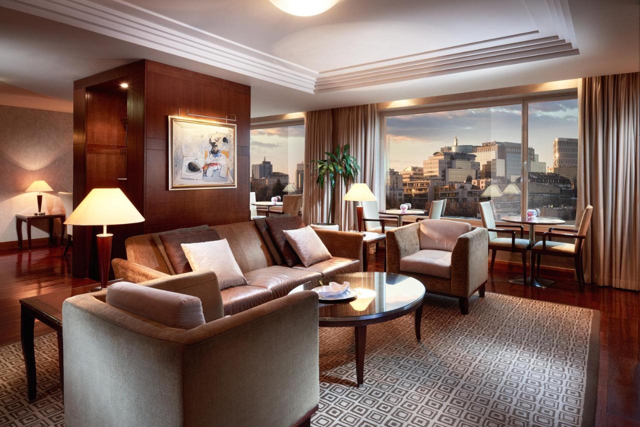 Regent Warsaw Hotel Buitenkant foto The image shows a stylish and modern living room with large windows offering a view of a city skyline. The room features a comfortable seating area with a sofa, armchairs, and a coffee table. There is a decorative piece of art on one wall, and soft l