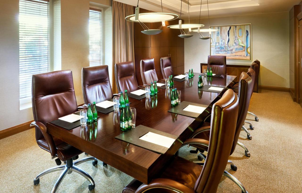 Regent Warsaw Hotel Buitenkant foto The photo shows a modern conference room with a long wooden table surrounded by several brown leather executive chairs. On the table, there are glass bottles of water and paper notepads, suggesting preparation for a meeting. The room features large w