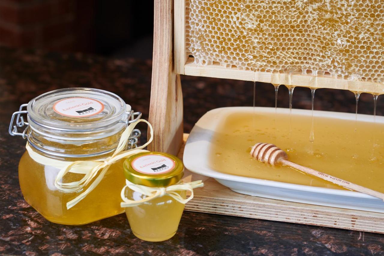 Regent Warsaw Hotel Buitenkant foto The photo shows a display of honey products. There is a honeycomb structure from which honey is dripping into a small plate. A wooden honey dipper rests on the plate, which contains liquid honey. Additionally, there are two jars of honey: one larger 