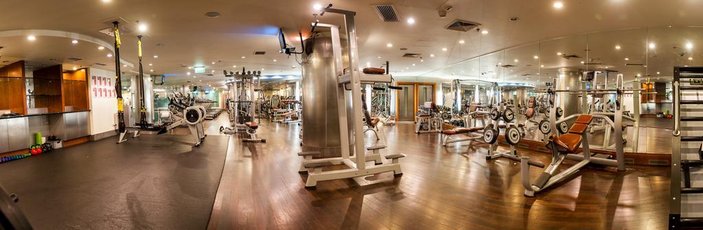 Regent Warsaw Hotel Buitenkant foto The photo shows an interior gym facility equipped with various exercise machines and weights. The space features a wooden floor and is well-lit, with overhead lighting and large mirrors on the walls. In the foreground, there is a multi-use exercise m